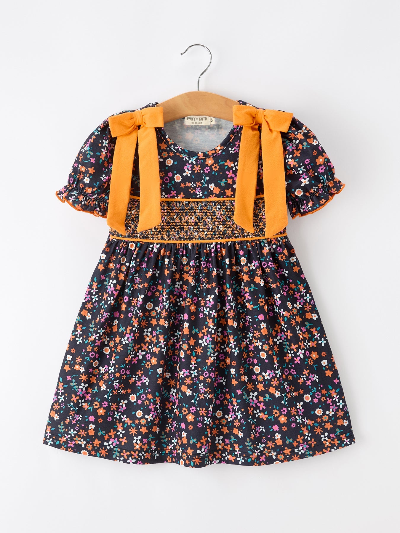 Girl dress with orange and floral print, smocked bodice, puff sleeves, orange bow shoulder details, and a flowy skirt.