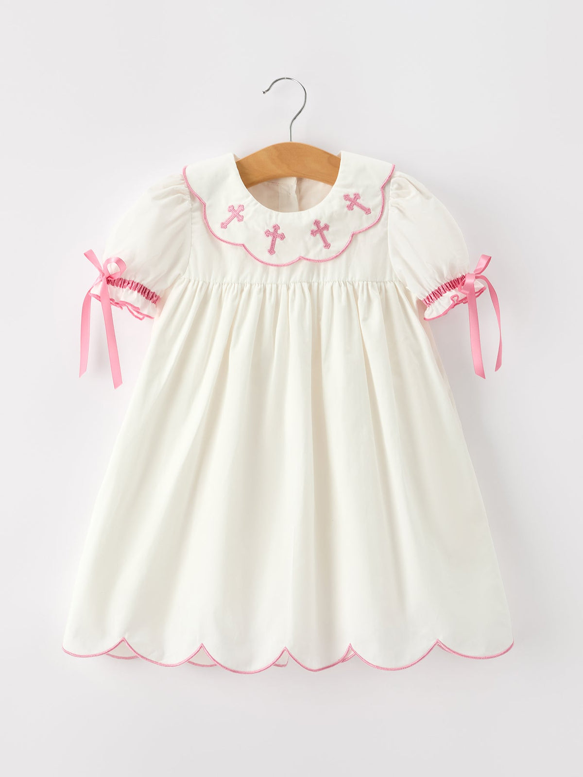 Ivory girls dress with pink cross embroidery, scalloped collar, puff sleeves, ribbon ties, and scalloped hem.