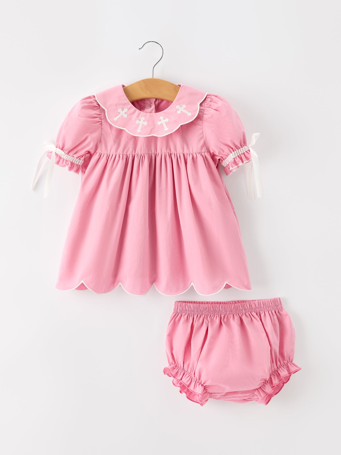 Pink christening bloomer set with white cross embroidery, scalloped collar, puff sleeves, ribbon ties, and matching bloomers.