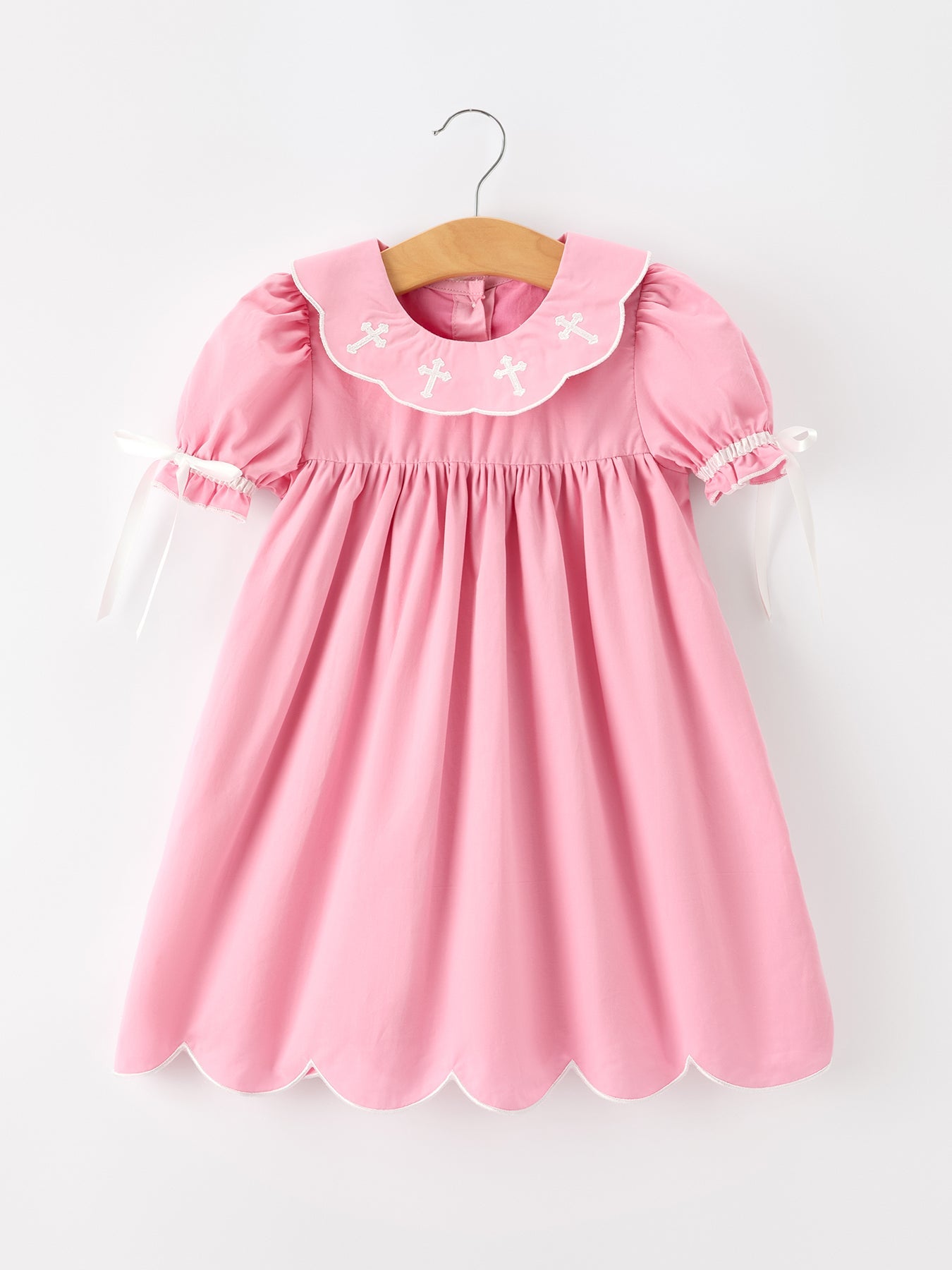 Pink christening dress with white cross embroidery, scalloped collar, puff sleeves, ribbon ties, and scalloped hem