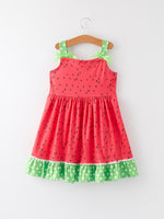 Smiling young girl wearing a pink and green watermelon print suspender dress by Rylee Faith Designs, playing outdoors.