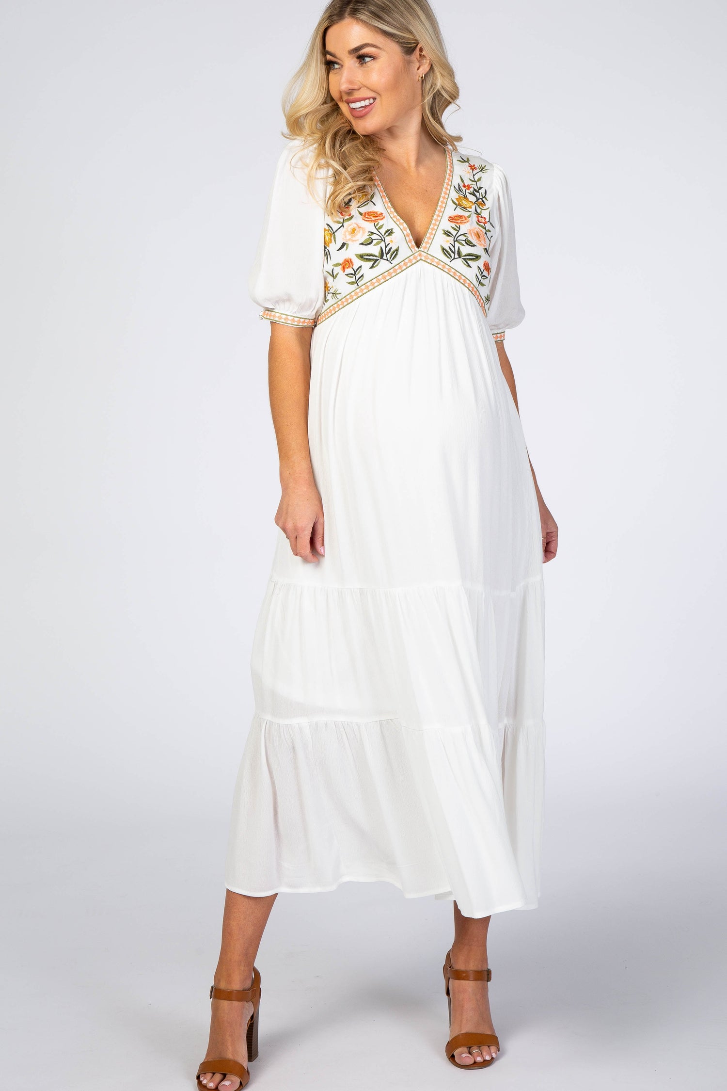 Ivory floral embroidered maternity midi dress with empire waist, tie straps, and midi length, worn with heeled sandals.