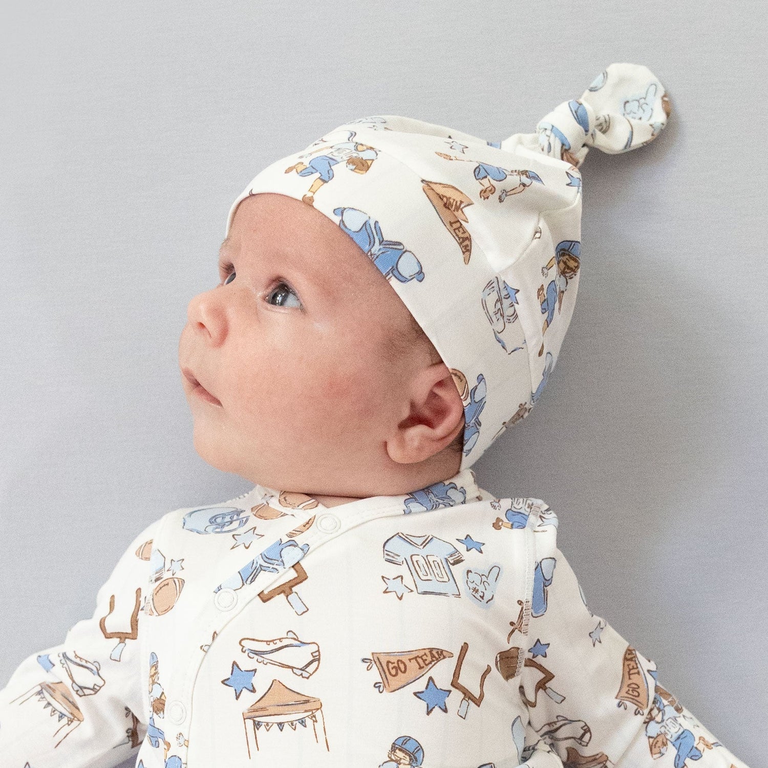 Knotted baby hat with a top knot in a football nursery print, made of soft stretchy fabric.