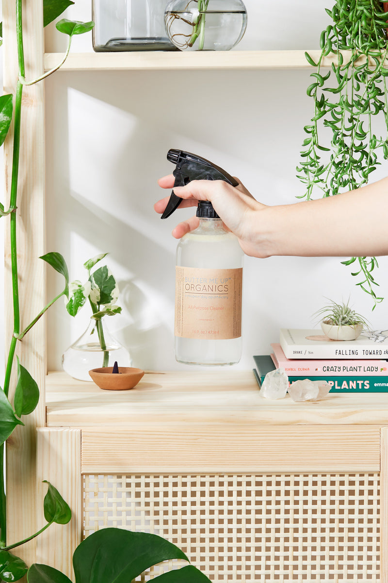 Natural all-purpose cleaner in a glass bottle with dual spray pump, surrounded by plants and books on a shelf.
