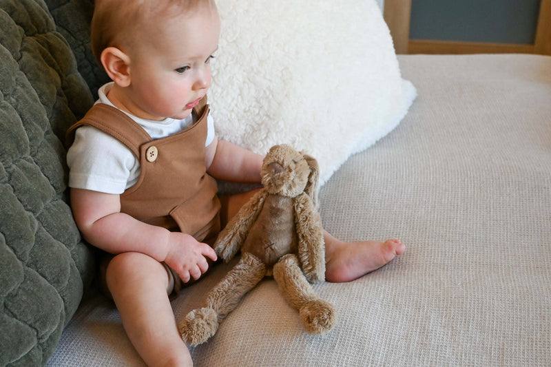 Baby holding Tiny Old Pink Rabbit Richie stuffed animal by Happy Horse on a sofa.