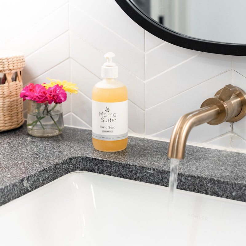 Hand soap bottle on bathroom counter with flowers, near running water faucet.