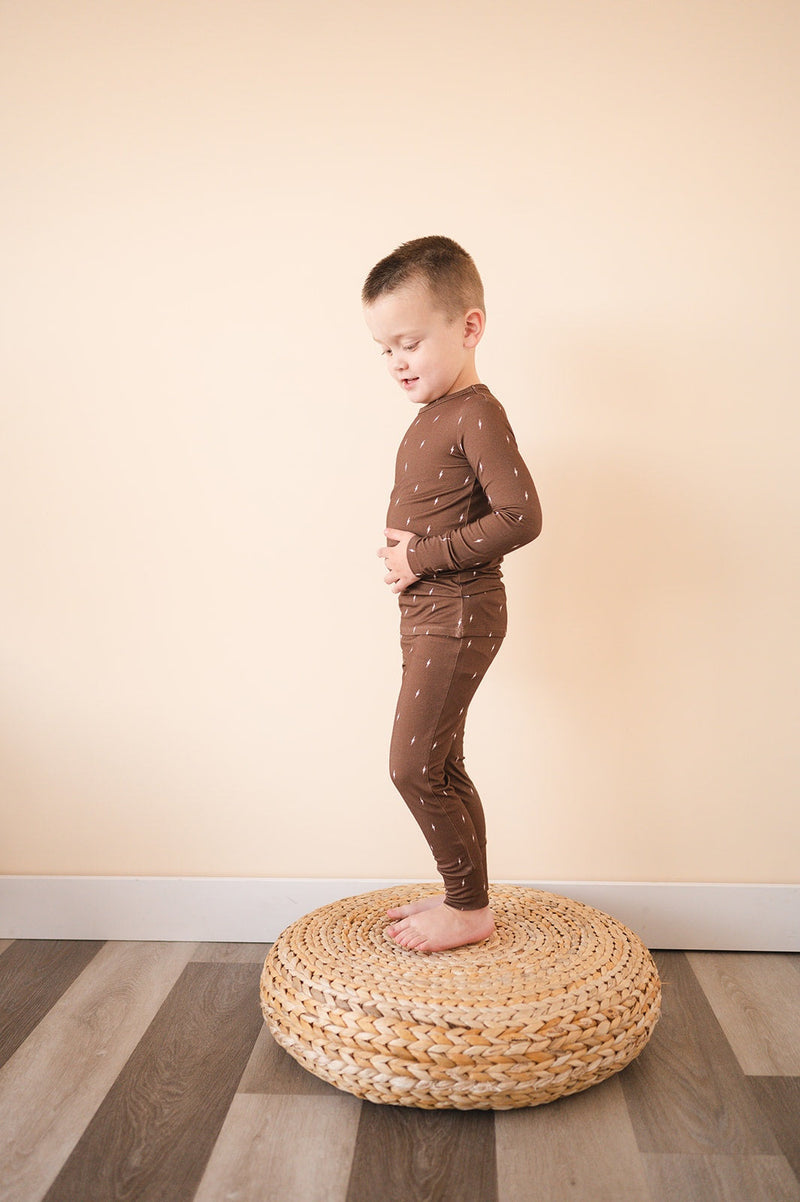Two-piece pajama set in brown with lightning pattern, featuring long sleeves and pants, worn by a child standing on a woven cushion.