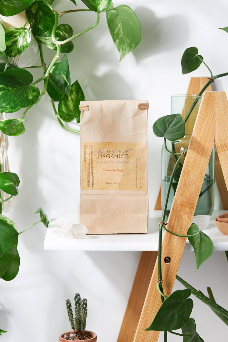 All Natural Dishwasher Soap by Butter Me Up Organics displayed on a shelf, surrounded by plants.