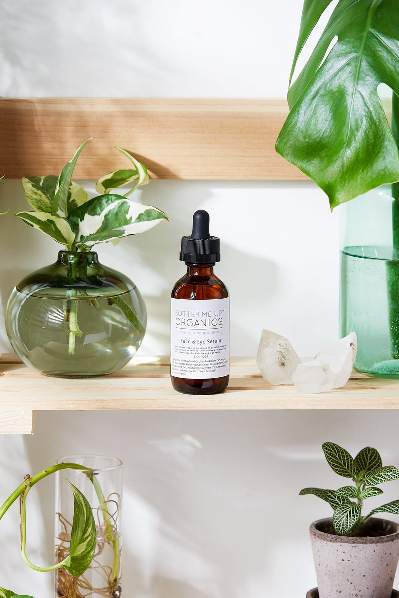 Organic Face & Eye Serum by Butter Me Up Organics shown on a wooden shelf surrounded by plants.