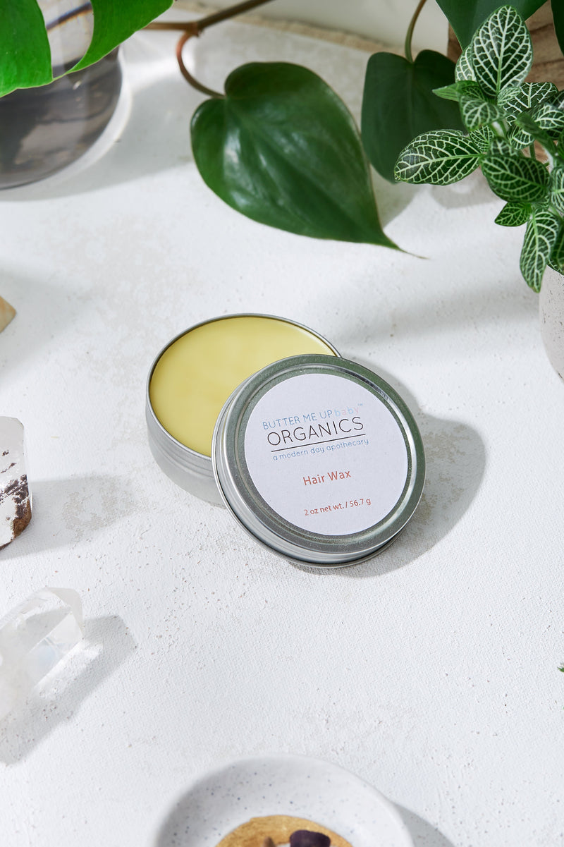 Organic Hair Wax in a tin container on a white surface surrounded by green plants.