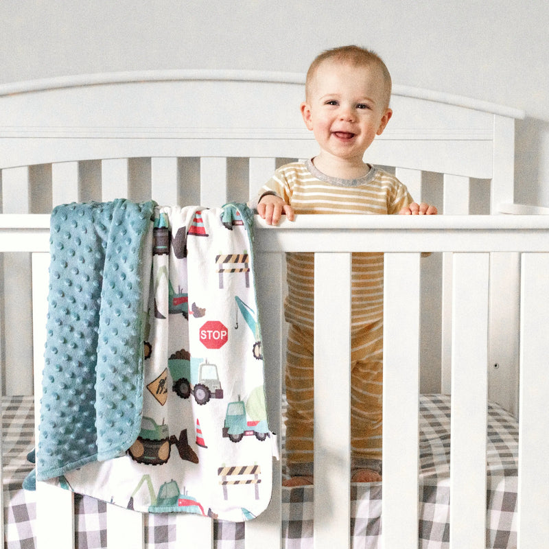 Baby standing in crib with construction trucks minky blanket, perfect for toddlers.