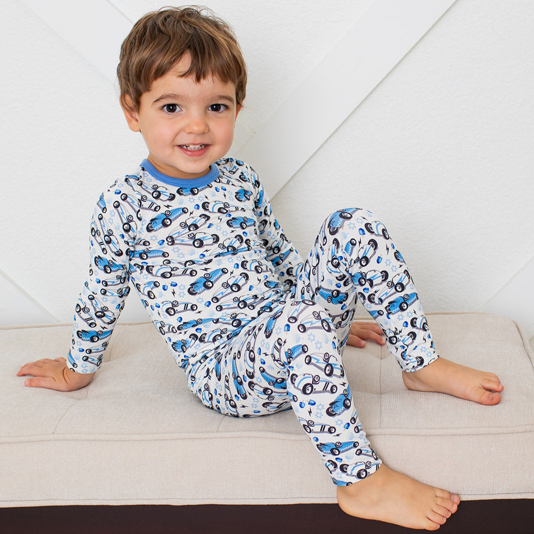 Long-sleeve baby pajamas with blue and white race cars, menorahs, Stars of David, and dreidels on a light background.