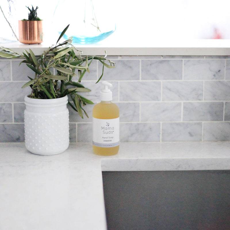Hand Soap in an 8oz glass bottle on a marble countertop with plants in the background.
