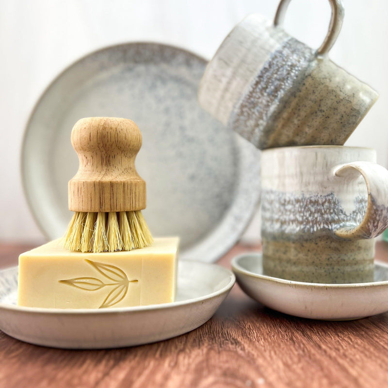 Solid dish soap bar with bamboo pot brush on ceramic dishes.