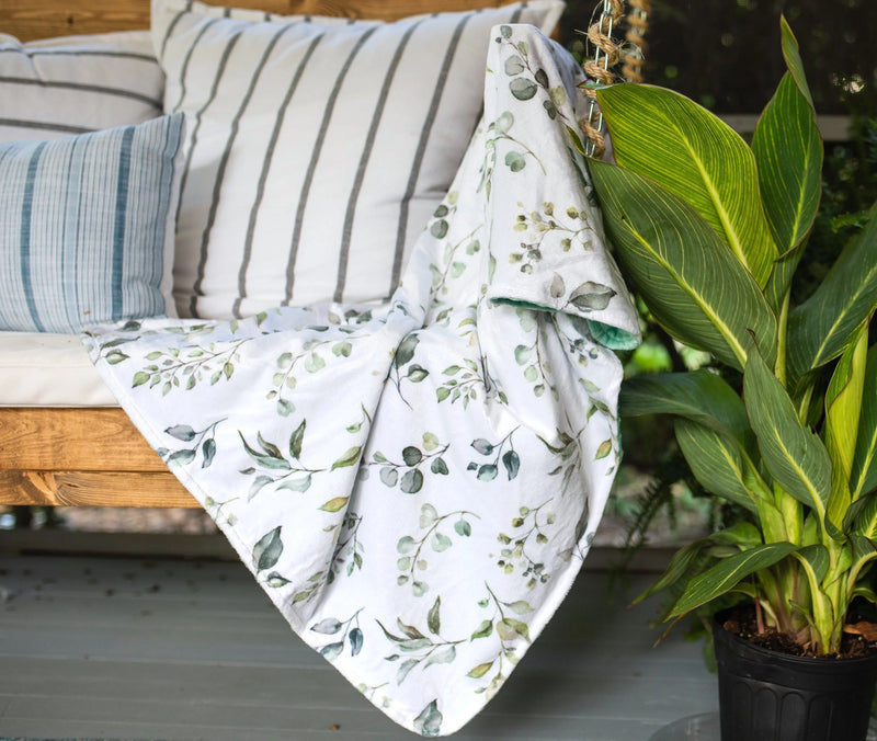 Premium baby and toddler minky blanket in eucalyptus greenery design, displayed on a cozy outdoor bench.