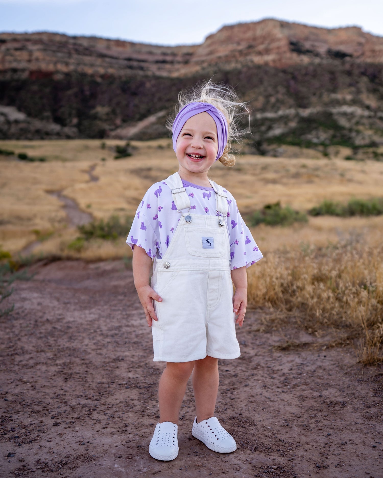 Ivory denim short overalls with adjustable shoulder straps and snap closures for a baby.