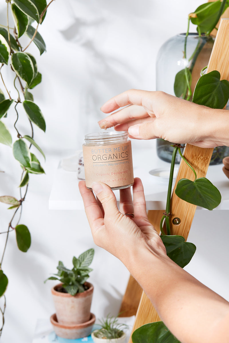 Organic deodorant in a clear glass jar being held in a plant-filled environment.