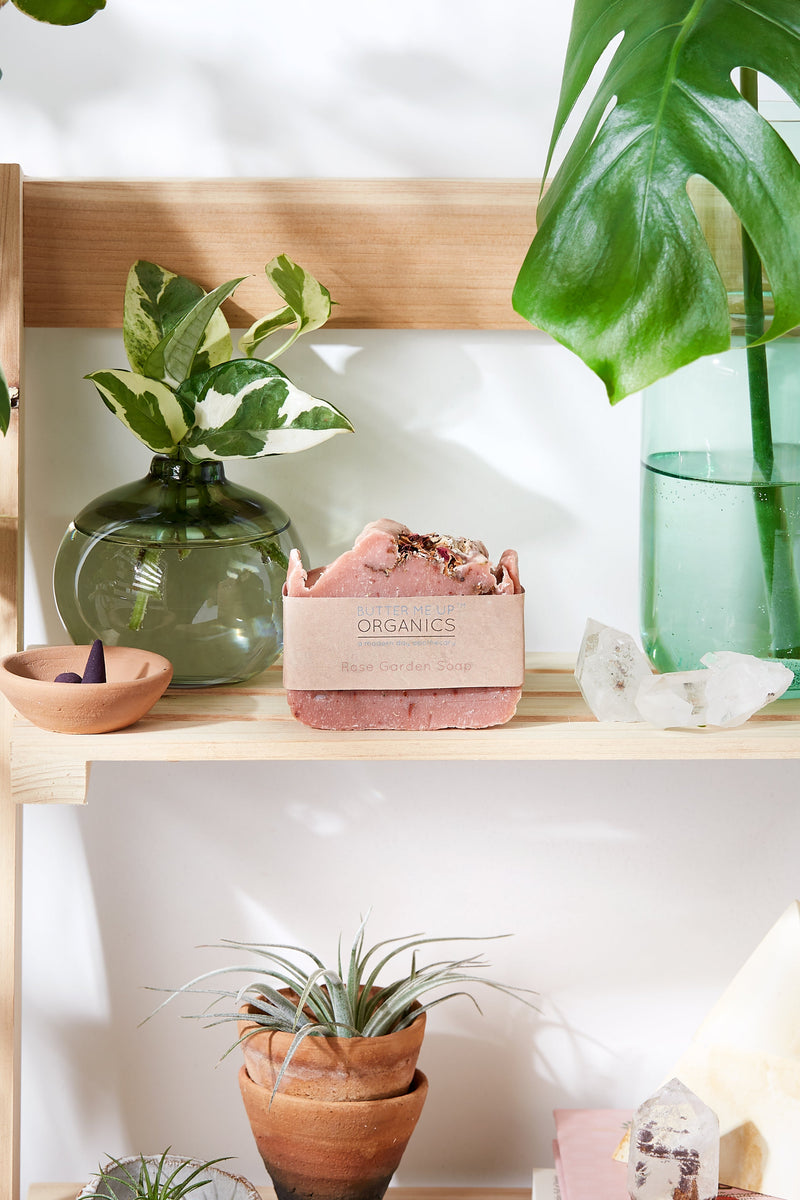 Rose Garden Organic Soap on wooden shelf with green plants and crystals, featuring natural ingredients and pink clay.