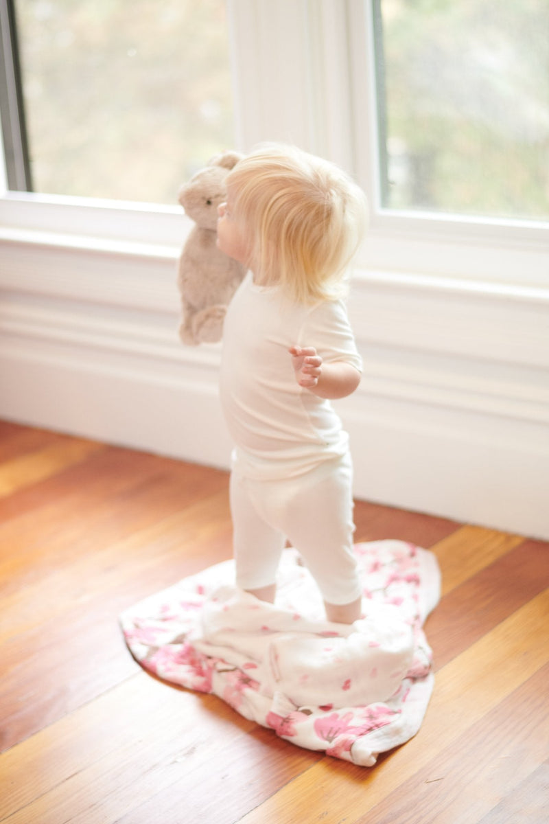 Toddler standing on Cherry Blossom Bamboo Muslin blanket near a window.