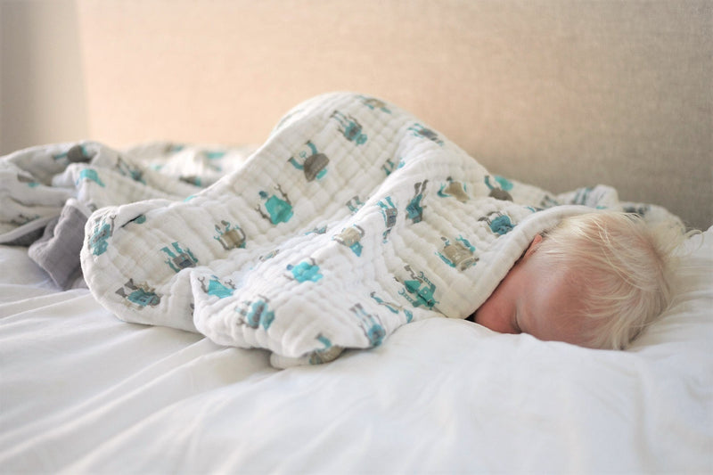Child sleeping under white robot-themed cotton muslin blanket on a bed.