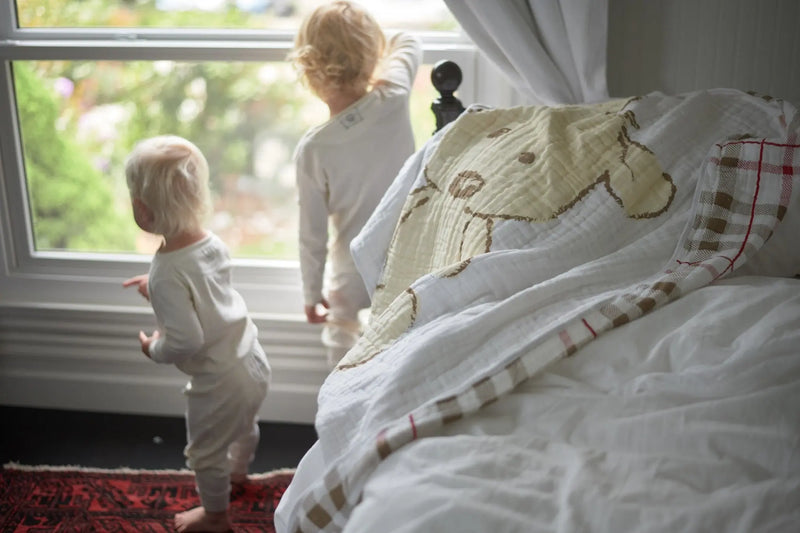 Teddy bear and plaid cotton muslin blanket on bed with two children by the window.