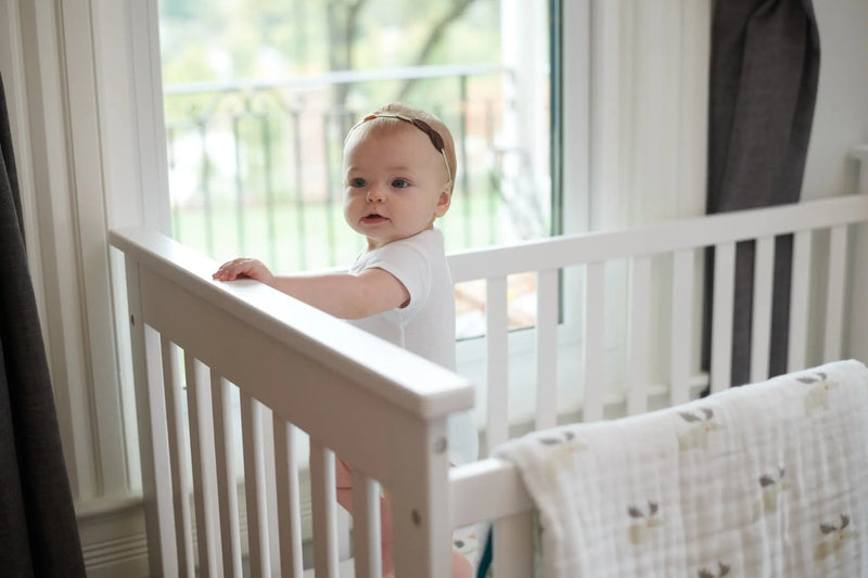 Baby in crib with Mister Moose and Forest Arrow Cotton Muslin Blanket draped over rail.