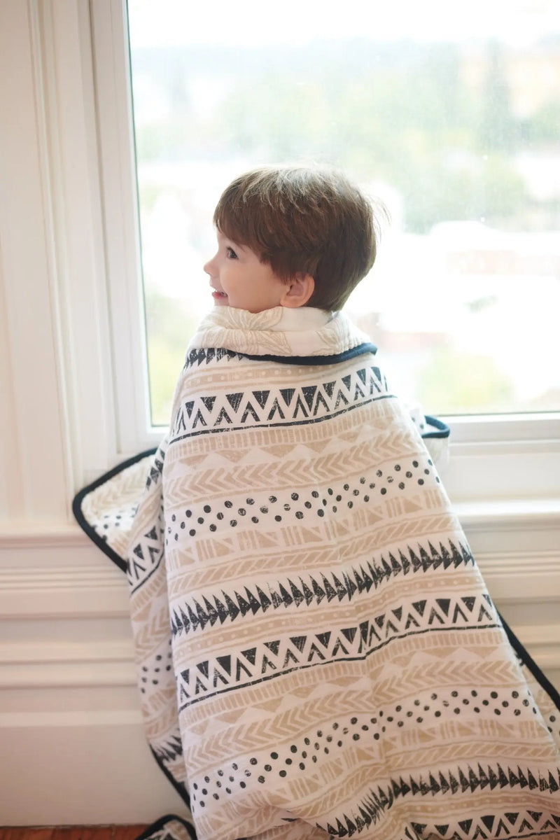 Child wrapped in Pyramid and Boho Fields cotton muslin blanket by a window.