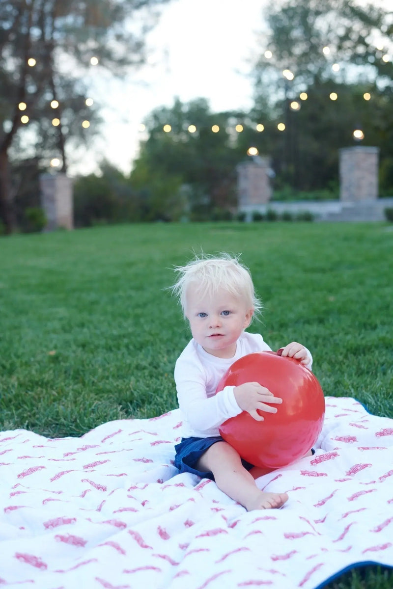 Child sitting on a cotton muslin blanket with fire truck and Dalmatian designs in a grassy area.