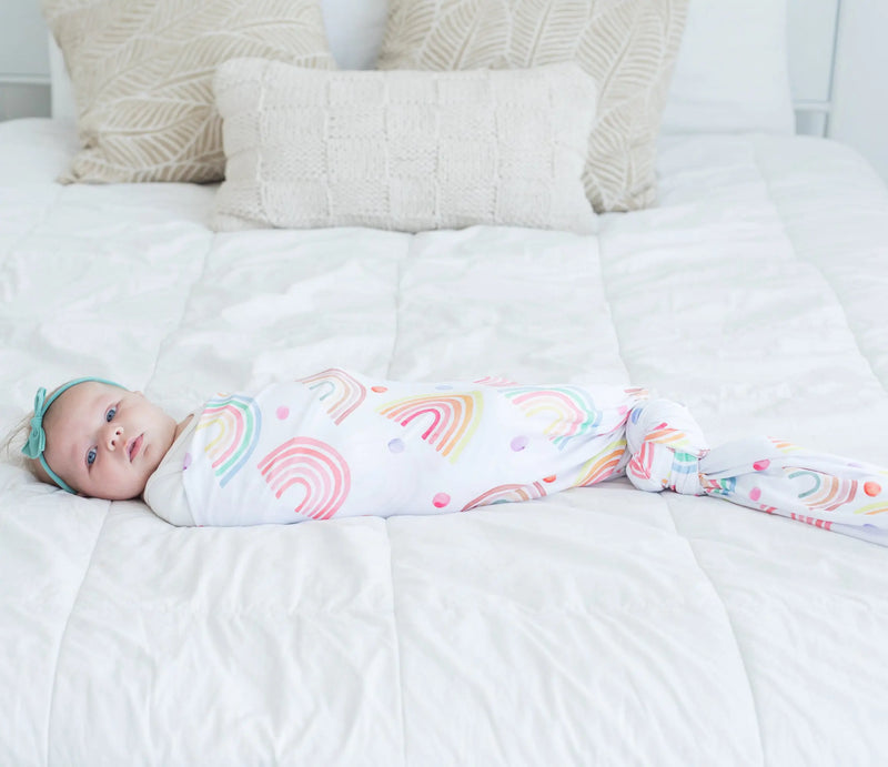 Buttery soft and stretchy swaddle blanket with rainbow pattern wrapped around a baby on a white bed.