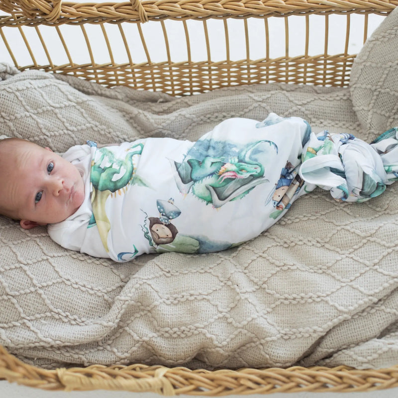buttery soft and stretchy swaddle blanket with dragon and knight print enveloping a baby in a woven basket
