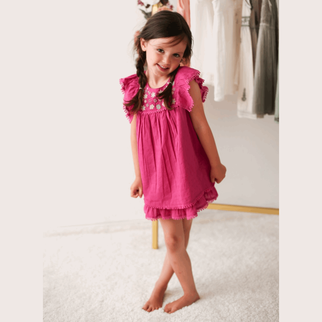 Hot pink mini dress with embroidery and pom pom details, worn by a young child with brown hair.