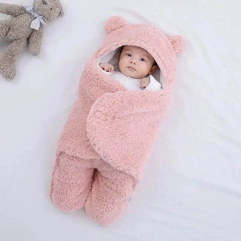 Baby in pink plush fleece sleeping bag with bear ears, lying on a white surface with a gray teddy bear nearby.