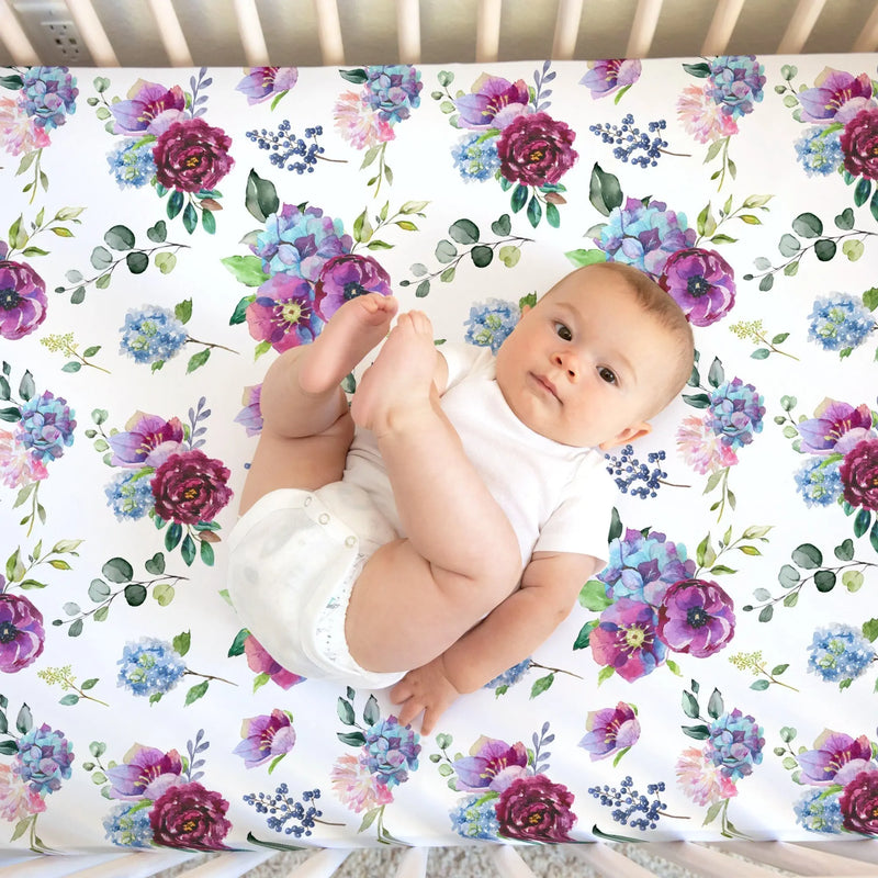 Blue floral fitted crib sheet with baby lying on soft fabric backdrop.
