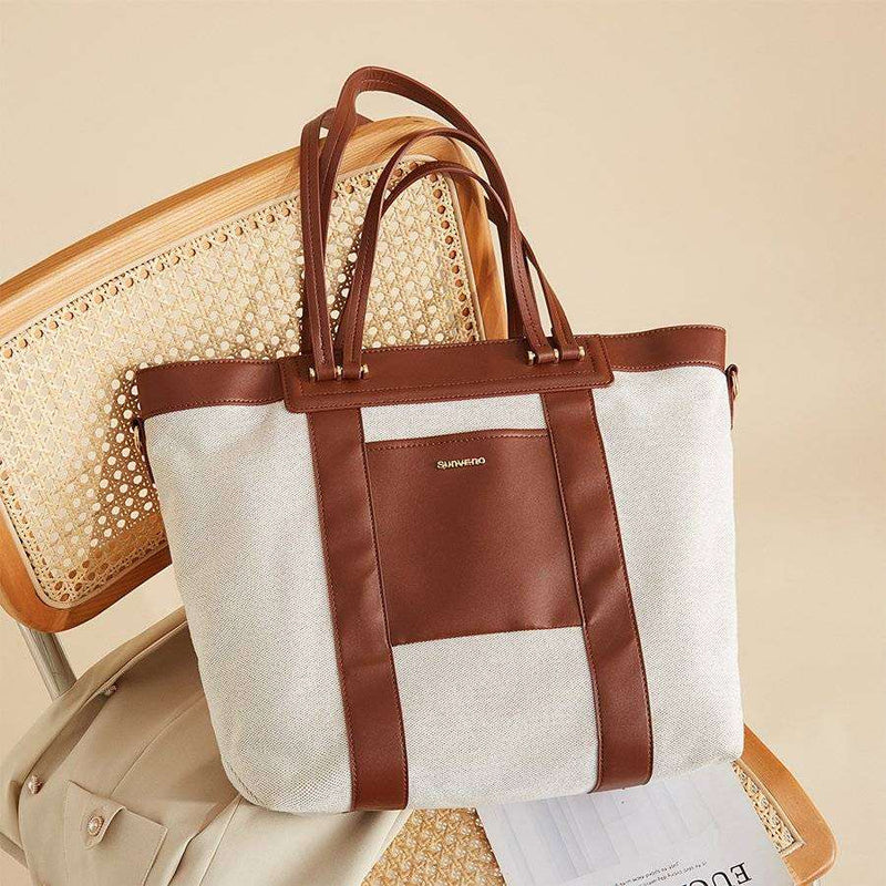 Canvas Tote Diaper Bag with brown accents, featuring reinforced handles and multiple carrying options, on a chair.