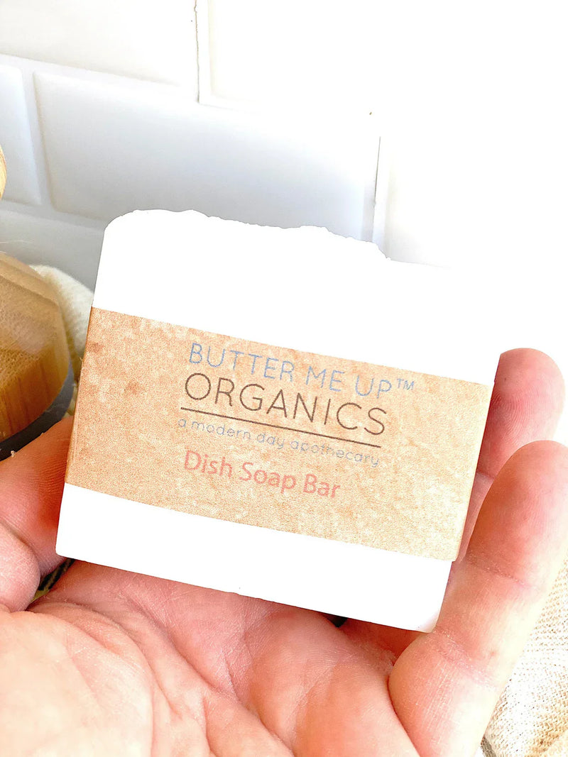 Organic solid dish soap bar held in hand, labeled "Butter Me Up Organics." Eco-friendly kitchen cleaning product.