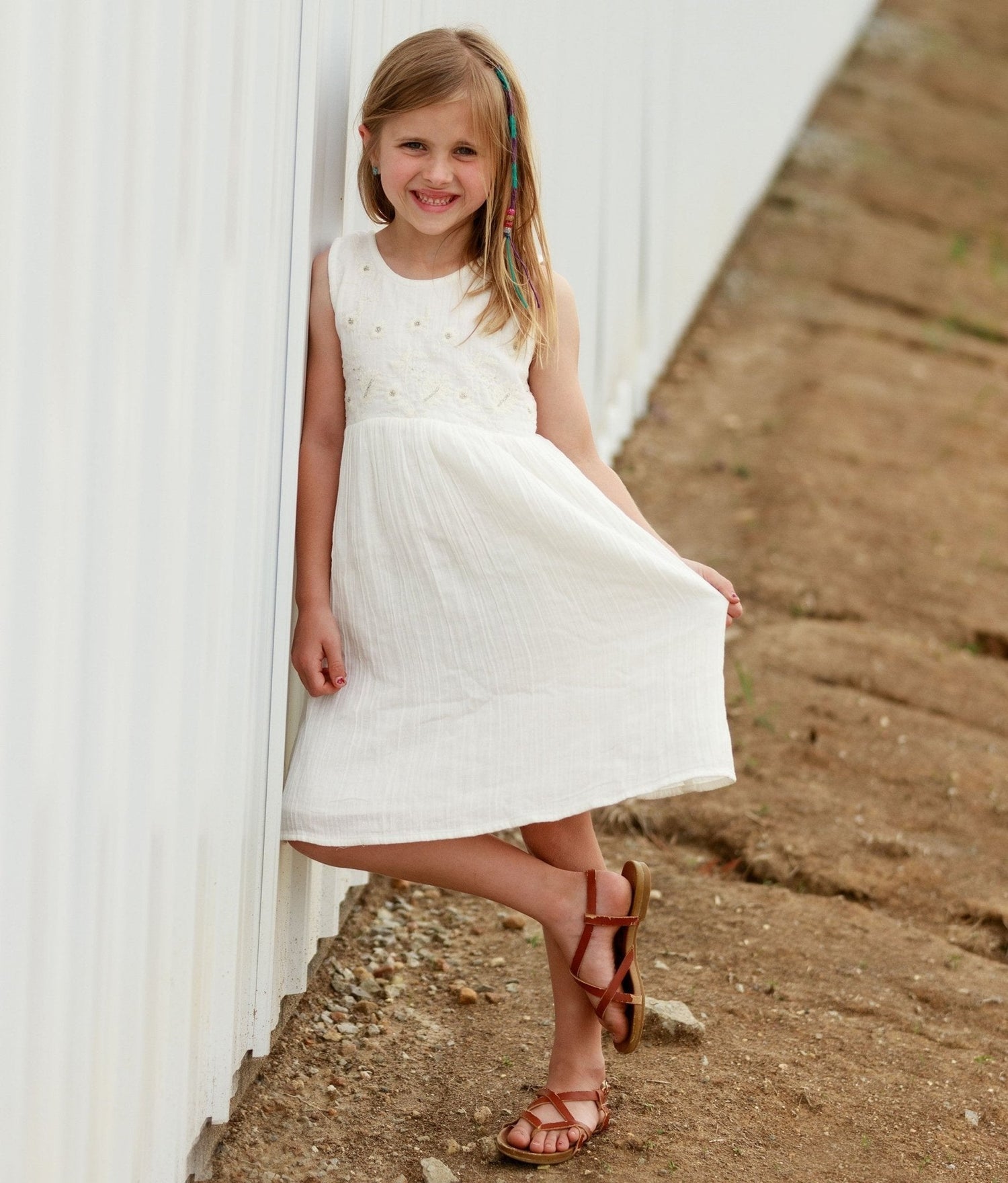 Off white baby dress with pleats, embroidery, and button closure, shown on a child with sandals.