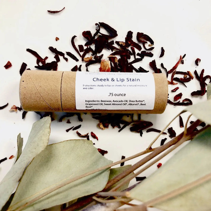 Organic natural cheek and lip stain in eco-friendly paperboard tube, surrounded by dried botanicals.