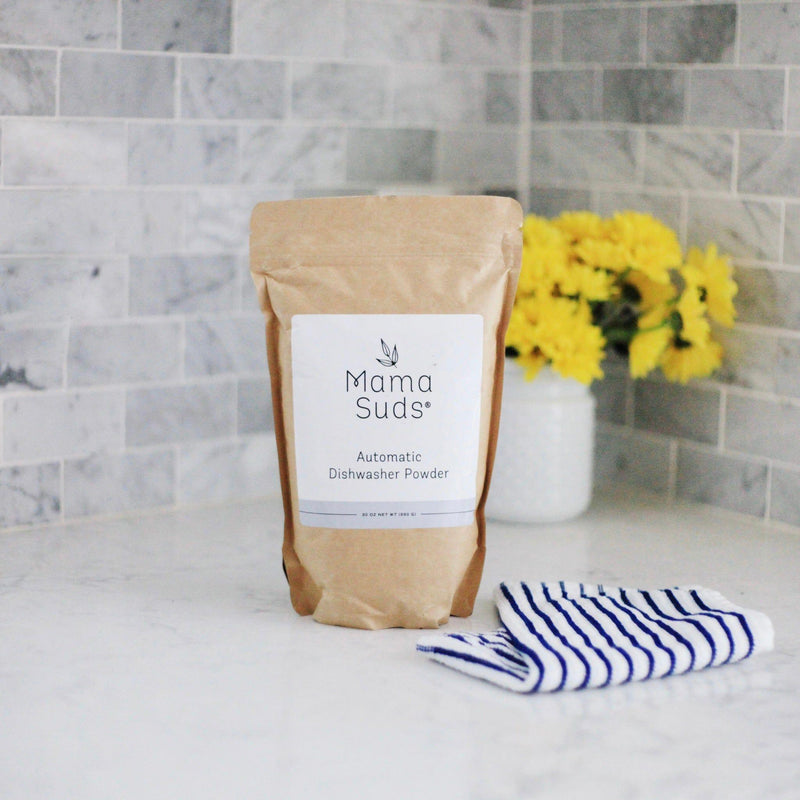 MamaSuds Automatic Dishwasher Powder in eco-friendly packaging on a kitchen countertop with a striped towel and yellow flowers.