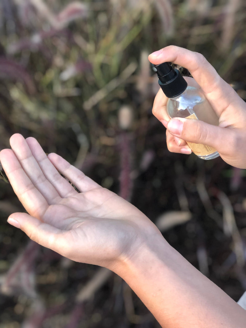 Organic hand sanitizer in glass spray bottle being applied to hands in an outdoor setting.