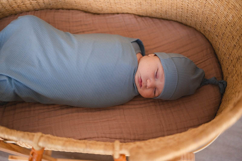 Newborn wearing a blue ribbed knot hat, swaddled in a matching blanket in a bassinet.