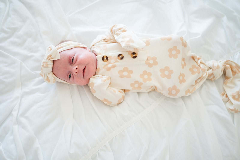 Knotted baby gown in blush daisy pattern featuring fold-over cuffs and a knotted bottom on a baby lying down.