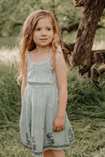 Sage green dress with a modern neckline, elastic waist, and embroidered detail at the hem, worn by a child.