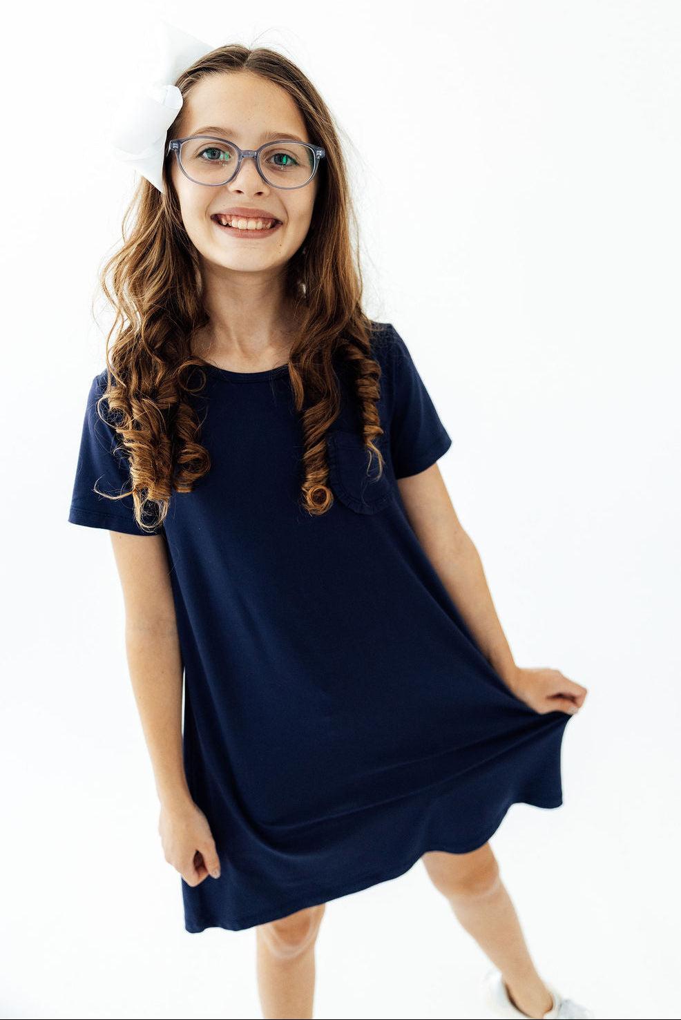 Navy short-sleeve t-shirt dress with a ruffle pocket, worn by a smiling girl with long hair.