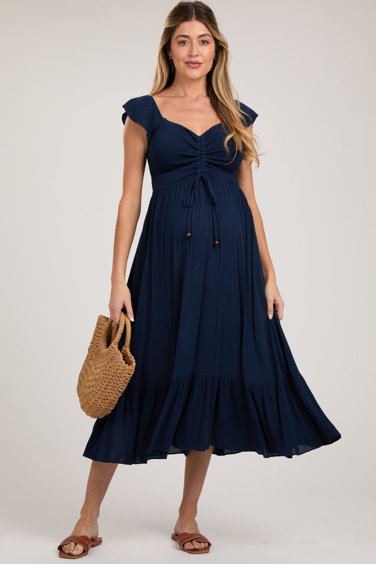 Pregnant model wearing a navy smocked ruched ruffle hem maternity maxi dress, smiling and holding her bump.