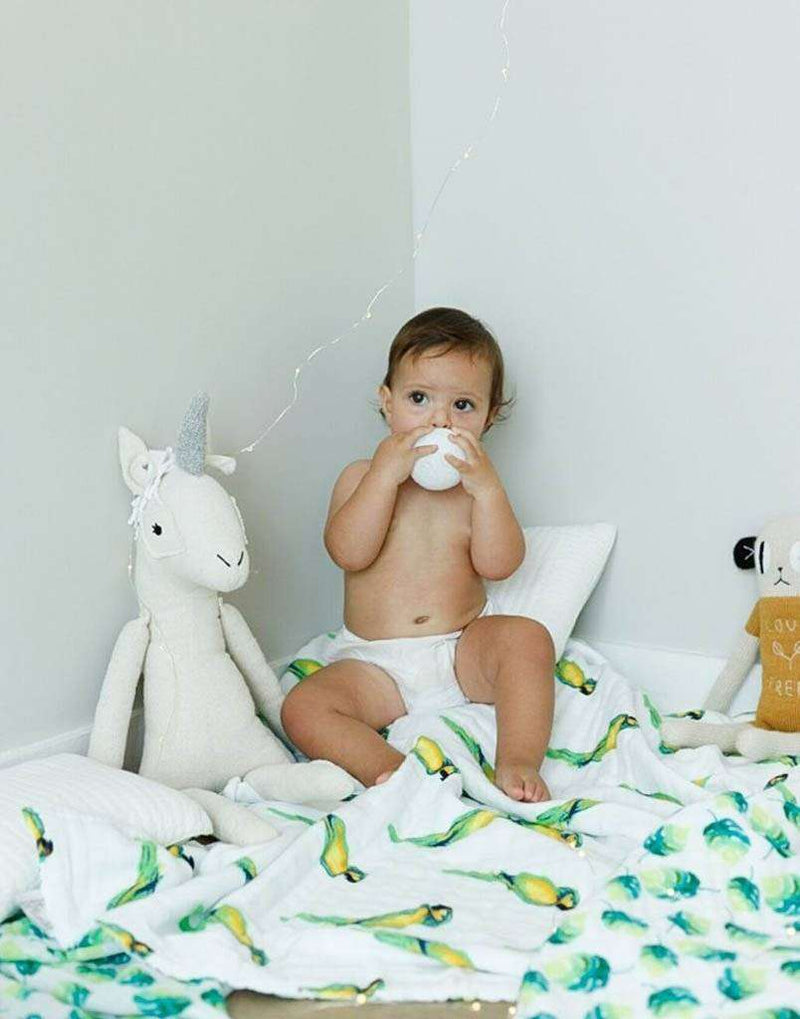 Baby sitting on Parrots Organic Snug Blanket with toy unicorn and stuffed animal.