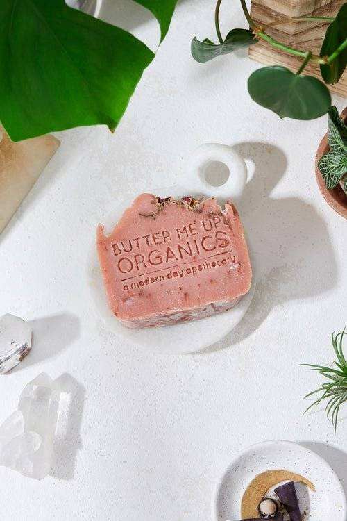 Rose Garden Organic Soap on a white dish, surrounded by plants, featuring an embossed design.