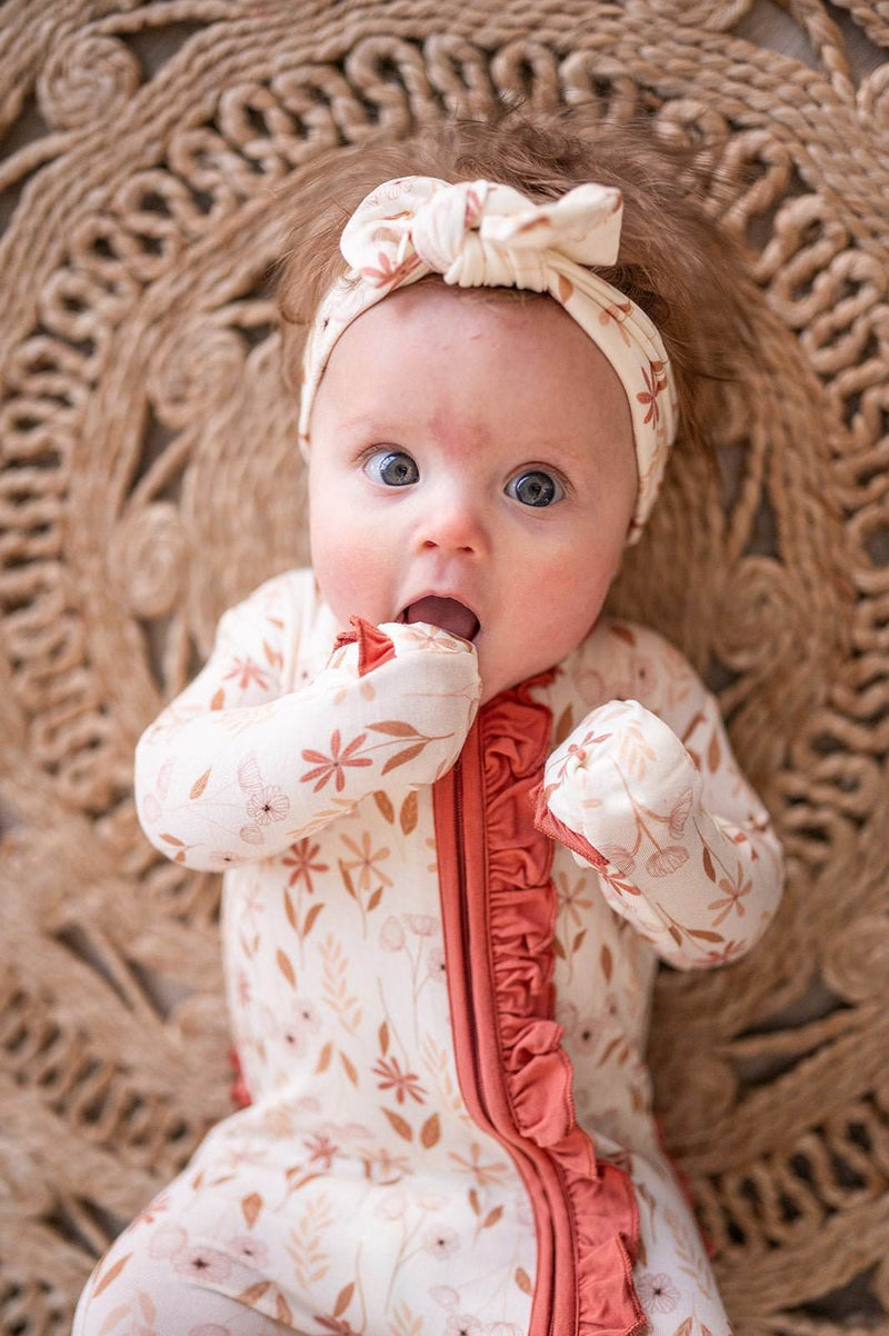 Baby in Wildflower Ruffle Zipper One Piece romper with floral design and ruffle details, laying on woven mat.