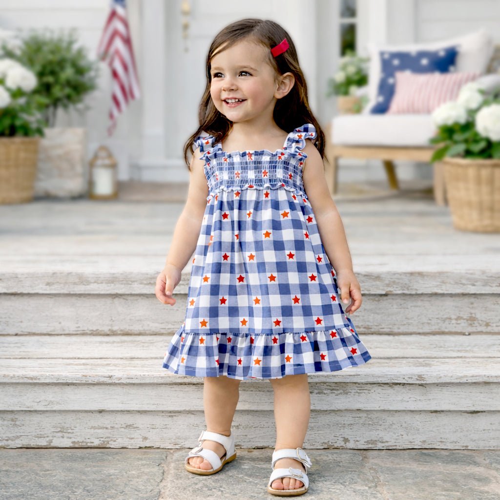 Blue gingham smocked dress with flutter sleeves and red star accents worn by a toddler girl with sandals.