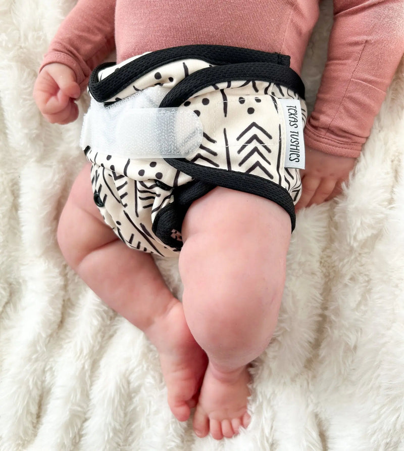 Newborn wearing Tiny Tushies Newborn Hook & Loop Cover with patterned design on a plush surface.