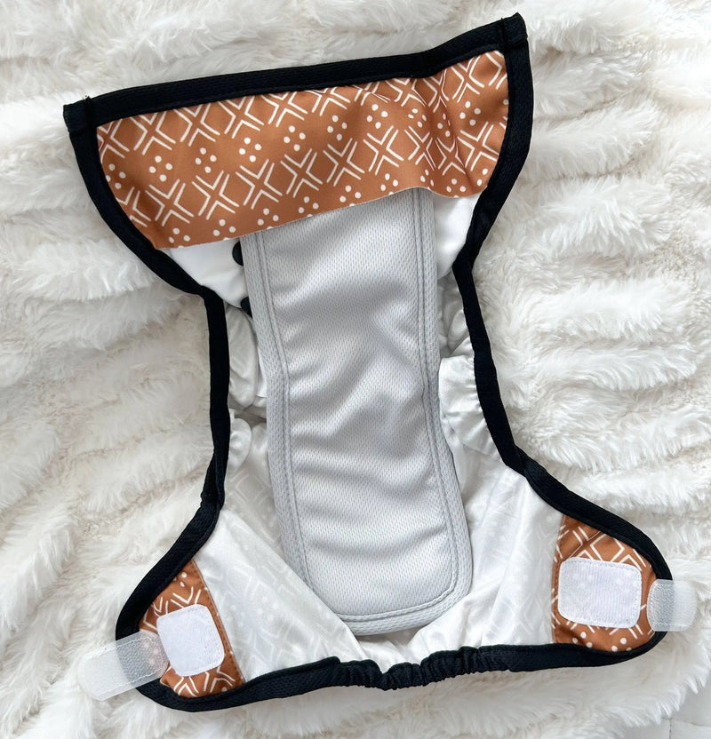 Tiny Tushies Newborn Hook & Loop Cover displayed open on a fluffy white surface, featuring adjustable rise settings and a wipeable interior for easy cleaning.